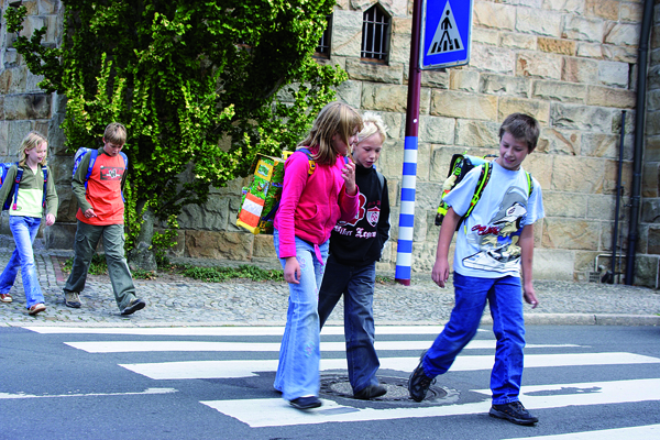 Mehrere Kinder überqueren einen Zebrastreifen vor einem steinernen Gebäude mit grünen Pflanzen. Alle tragen Schulrucksäcke. Ein Verkehrsschild mit Zebrastreifen-Symbol ist sichtbar. Das Bild vermittelt eine Botschaft der Sicherheit und Prävention im Straßenverkehr, passend zum Kontext der Unfallkasse NRW und ihrer Bemühungen um Arbeitsschutz und Unfallverhütung.