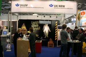 Ein Messestand der DGUV und Unfallkasse NRW auf einer Messe. Der Stand zeigt Schilder mit der Aufschrift 'Lass dich sehen!' und 'Risiko raus'. Zwei Frauen sprechen miteinander, weitere Besucher sind um den Stand gruppiert. Das Thema des Standes fokussiert auf Arbeitsschutz und Prävention gegen Unfälle bei der Arbeit.