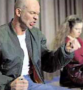 Eine kahle Person in einer olivgrünen Jacke sitzt links auf einer Bühne und spricht gestikulierend. Eine zweite Person im Hintergrund mit langen, braunen Haaren und einem roten Pullover ist ebenfalls zu sehen. Die Szene könnte im Kontext von Arbeitsschutz und Prävention bei der Unfallkasse NRW verwendet werden, um Kommunikation und Interaktion zu verdeutlichen.