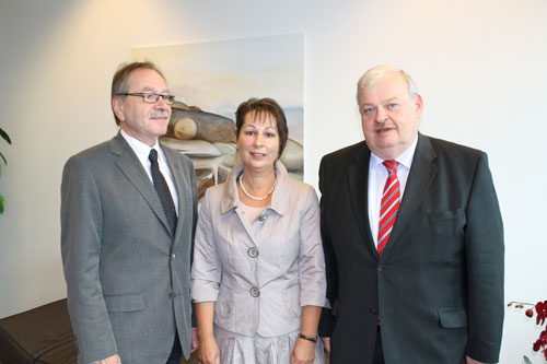 Drei Personen, zwei Männer und eine Frau, in formeller Kleidung stehen vor einem Gemälde. Die Szene scheint in einem Büroumfeld stattzufinden, was auf ein geschäftliches oder formales Treffen hinweist. Der Kontext könnte im Bereich der Unfallprävention und Arbeitssicherheit angesiedelt sein, passend zur Unfallkasse NRW.