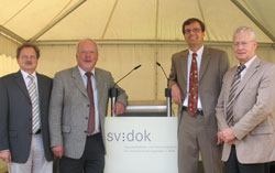 Vier Männer stehen vor einem Rednerpult in einem Zelt. Das Rednerpult ist mit einem Logo beschriftet. Die Männer tragen Anzüge und scheinen in einer formellen Veranstaltung zu sein, die möglicherweise der Arbeitsschutz- oder Präventionsarbeit der Unfallkasse NRW gewidmet ist. Im Hintergrund sind die Zeltwände zu erkennen.