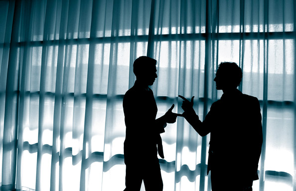 Zwei Personen stehen in Silhouettenform vor bodenlangen Vorhängen und diskutieren mit einander. Die Szene ist in einem Büro- oder Konferenzraum angesiedelt und symbolisiert einen Austausch oder eine Auseinandersetzung, die im Rahmen von Arbeitsschutz oder Prävention relevant sein könnte. Die Personen sind nicht identifizierbar, was den Fokus auf das Thema Kommunikation und Problemlösung legt. Der Raum hat ein weiches, gleichmäßiges Licht, das durch die Vorhänge gefiltert wird, was eine professionelle Atmosphäre unterstreicht.